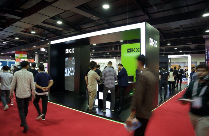 The image shows a bustling trade show floor with attendees gathered around the OKX booth, featuring prominent green and white branding displaying "OKX" logos. The scene aligns with the post, capturing the lively Erbil event where visitors can meet the OKX team to learn about their crypto exchange platform.