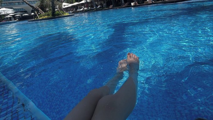 The image shows a person's feet floating above water at a swimming pool, with their legs spread out as they enjoy the water. The photo appears to have been taken from an overhead perspective or a low angle, giving the impression that the person is in mid-air while still being in the water. This creates a sense of weightlessness and relaxation. The image also includes text that reads "gm good morning small steps every day still move you forward stay consistent even when motivation feels low."
