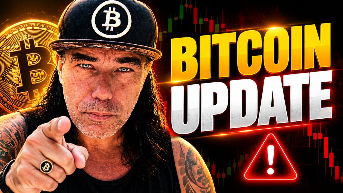 A long-haired man wearing a Bitcoin-branded cap and ring points directly at the camera, set against a backdrop featuring a large Bitcoin logo, candlestick price charts, and bold "BITCOIN UPDATE" text alongside a red warning triangle emphasizing the urgent message.