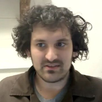 A close-up photo of a man with curly dark hair, a mustache, and a short beard, wearing a brown jacket over a gray shirt, with a neutral indoor background. No text or numbers are visible in the image.