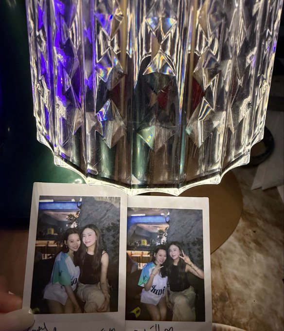 A polaroid photo held in front of a decorative cut-glass lamp, showing two young women posing together in two frames, one making a peace sign.