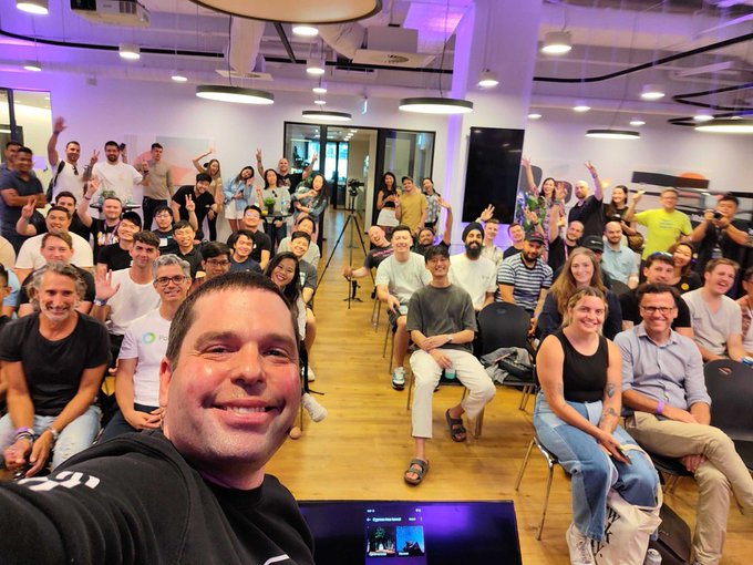 A selfie taken by a smiling man in a black hoodie at what appears to be a tech meetup or office event, with a large crowd of people behind him posing and waving in a modern venue with purple accent lighting.