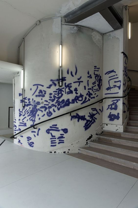 A stairwell corner is covered in blue spray-painted graffiti that includes Chinese characters, abstract symbols, and the letters "a/d." A metal handrail runs along the painted walls, illuminated by vertical fluorescent light fixtures.