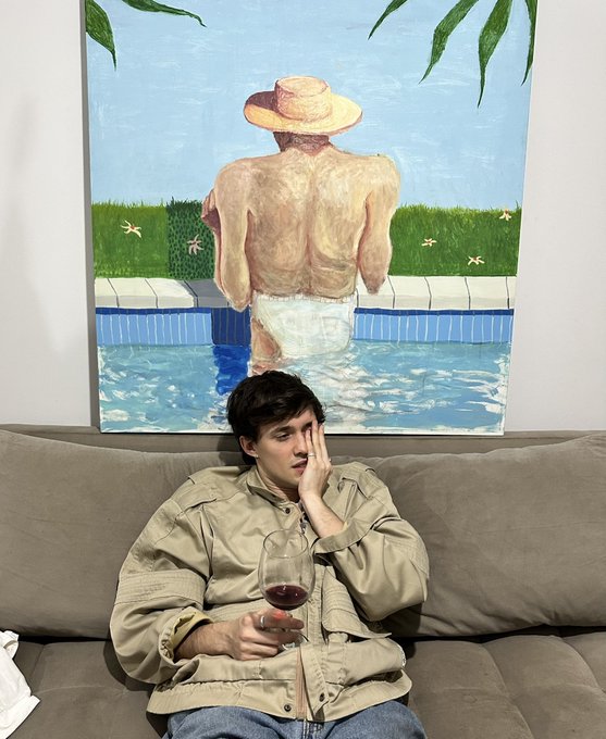 A young man sits on a brown couch holding a glass of red wine, appearing bored, with a painting of a person in a pool hanging on the wall behind him.