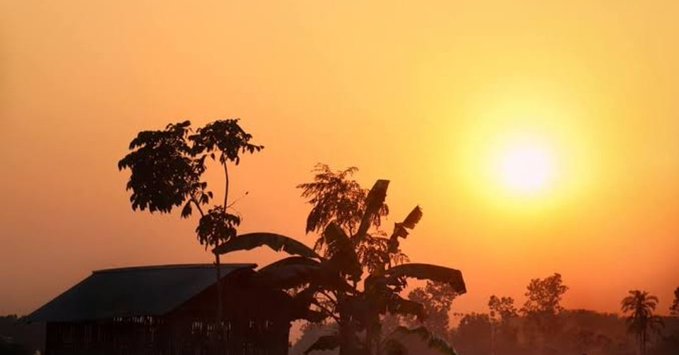 A silhouette of trees and a small building against a hazy, golden-orange sky as the sun sets.