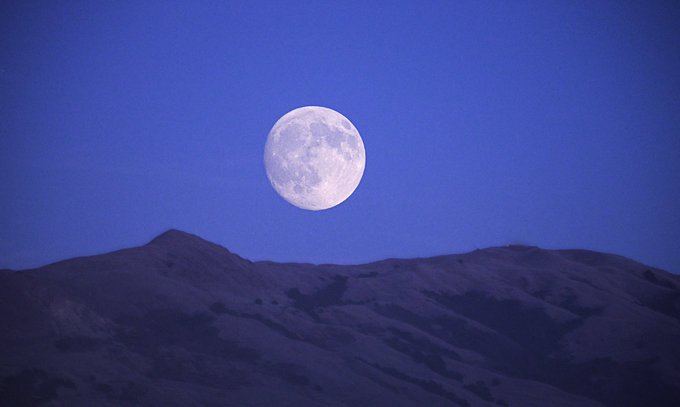 A large, bright full moon hangs in a deep blue twilight sky above the dark, silhouetted ridgeline of a mountain range.