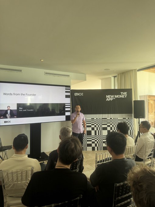 A man speaks at the OKX Creator Retreat in Ibiza, standing before a screen displaying "Words from the Founder" with Star Xu, CEO and Founder, alongside banners reading "OKX" and "THE NEW MONEY APP," to an attentive seated audience.