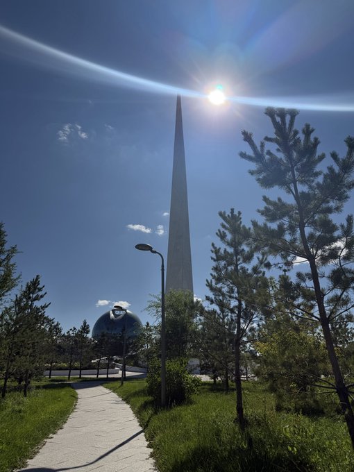 A sunny outdoor view of a tall, thin monument (Bayterek Tower) and a golden spherical structure set within a park with pine trees and a walking path.