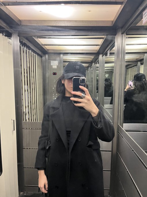 A mirror selfie of a person wearing an all-black outfit with a long coat and a black baseball cap, taken in what appears to be an elevator.