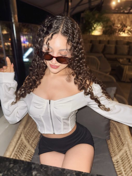 A woman with curly dark hair and sunglasses wears a white off-the-shoulder corset top and black shorts, sitting in an outdoor lounge area.