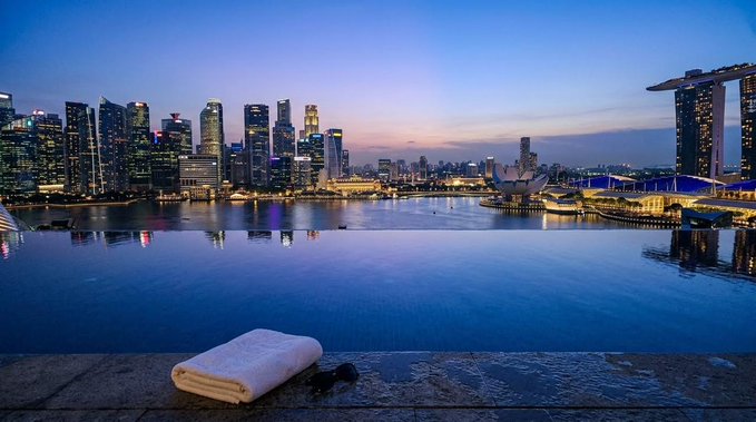 A folded white towel rests on a stone ledge overlooking Marina Bay at twilight, with the illuminated Singapore skyline—including the iconic Marina Bay Sands—glowing across the calm water. The serene luxury setting visually reinforces the post's pitch for discreet, frictionless hotel bookings at premier crypto conferences like Token2049.