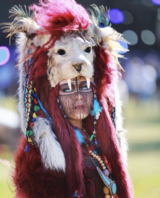 A person wearing an elaborate ceremonial headdress featuring a bleached animal skull with fangs, vibrant pink and red feathers, long crimson faux fur, and intricate beaded jewelry with turquoise stones.