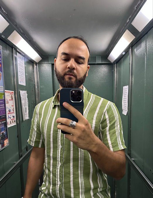 In the image, there's a man taking a selfie inside an elevator with his phone held up at the top of his head, looking down towards himself. The surrounding walls are painted green and blue, adding a vibrant touch to the scene. There is also a poster hanging on the wall next to him. The caption of the post reads: "Day 42 of posting selfie until $chog hits $2.1M". This suggests that the man might have been taking photos inside the elevator for several days in a row, possibly due to an ongoing challenge or competition involving selfies and their monetary values.