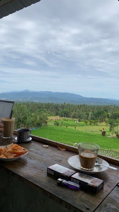 A scenic view from a cafe or restaurant window showing coffee drinks, snacks, and cigarettes on a wooden ledge overlooking lush green rice fields and distant mountains under a cloudy sky.