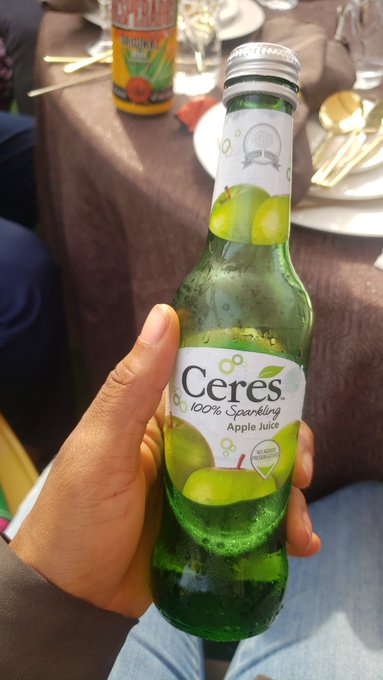 A hand holding a glass bottle of "Ceres 100% Sparkling Apple Juice" at a table set with plates and cutlery, with another beverage visible in the background.
