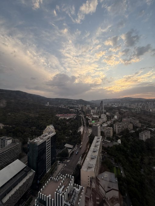 An aerial cityscape view at sunset, featuring modern high-rise buildings, tree-lined streets, and rolling hills beneath a dramatic sky filled with scattered clouds illuminated by golden light.