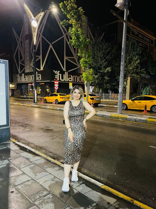 A nighttime street photo of a smiling woman in a leopard-print dress and white sneakers posing on a sidewalk, with the 'full moda' venue and yellow taxis visible in the background.