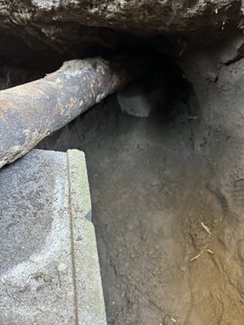 A view into a dark, narrow underground tunnel or culvert reveals exposed pipes running along the rocky ceiling above muddy ground.