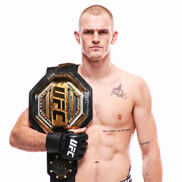A shirtless male UFC fighter with short blonde hair holds a UFC championship belt, with "UFC" and "Champion" visible on the gold plate, while wearing black "UFC" gloves and displaying tattoos on his chest and arms. The image represents Ian Garry, the fighter mentioned in the betting post, who is being wagered on to win against Islam Makhachev.