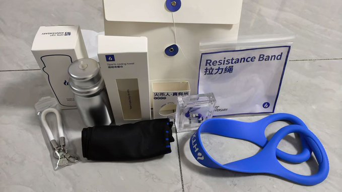 A collection of branded merchandise is displayed on a tiled floor, including a silver sports water bottle, a sports cooling towel, a blue resistance band with 'Lazada' branding, and other boxed items.