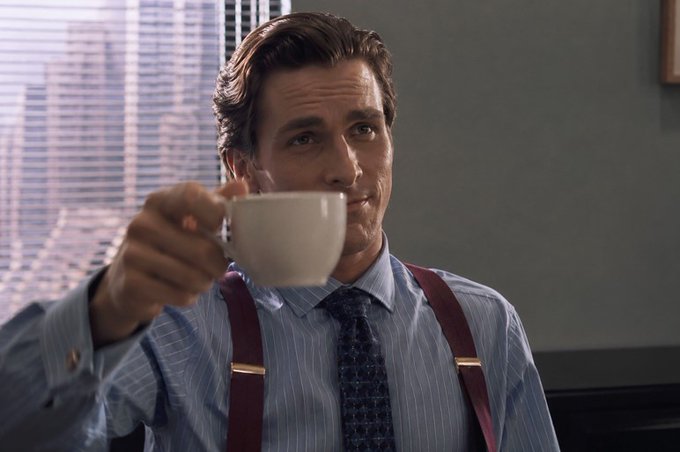 The image features a man sitting at a desk, holding a cup of coffee while wearing suspenders and a blue shirt with a tie. The background shows an office setting with a window that provides natural light. The caption for the image reads "good morning fam interesting update from @NomismaNetwork today, Nomisen ID mint cost is now tied to your wallet score across EVM ecosystems the more active your wallet the better your position also tapped my @useTria card this morning like it is just how i pay for things now".