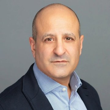 A headshot portrait of a middle-aged man with a shaved head, wearing a dark blazer over a light blue striped collared shirt, photographed against a neutral gray background. The image appears to be a professional headshot of Joseph Chalom, the Sharplink CEO mentioned in the accompanying post criticizing EIP-8363's potential impact on DeFi and institutional adoption.