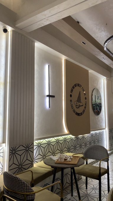An interior shot of a stylish café or restaurant corner with a small round table set for two, a wall-mounted vertical LED light, a circular mirror, decorative geometric tile patterns, and houndstooth-patterned chairs.