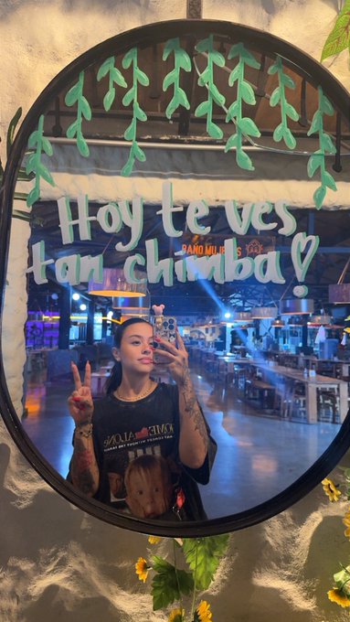 A mirror selfie of a young woman holding up a peace sign, with hand-painted green vines on the mirror frame and the Spanish text 'Hoy te ves tan chimba' with a heart symbol.
