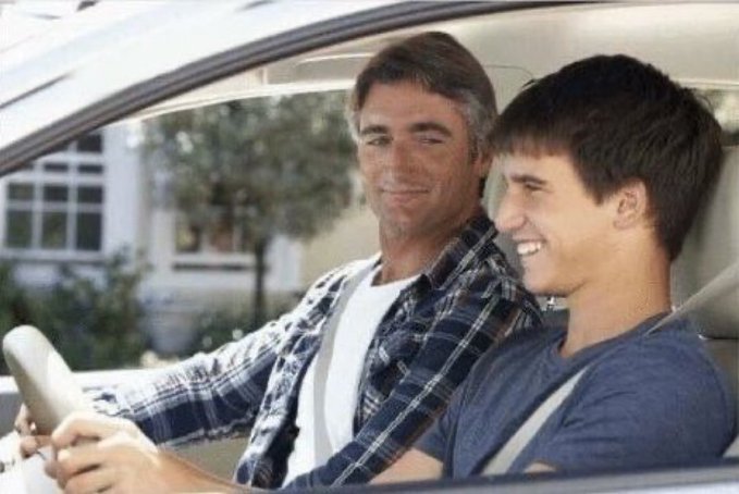 An older man with gray hair in a plaid shirt sits in the passenger seat of a car next to a smiling young man at the wheel.