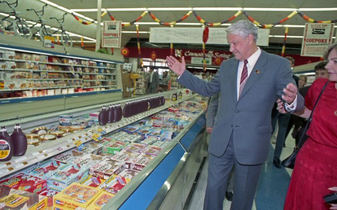 In a grocery store's deli section, an older man dressed in a suit stands next to a counter displaying an assortment of food items including cakes and pastries. A woman wearing red dress is walking by him. The people are surrounded by several large bins filled with various items, and a sign suggests that it might be the first time for them to experience hyperliquid trading as an alternative to traditional foods.