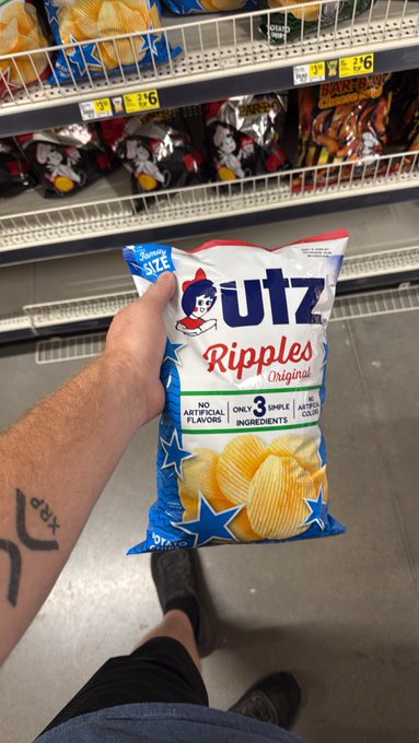 A hand holds up a bag of Utz Ripples Original potato chips in a grocery store aisle, showing the patriotic red, white, and blue packaging with text highlighting "NO ARTIFICIAL FLAVORS" and "ONLY 3 SIMPLE INGREDIENTS," playing on the visual resemblance to the XRP ($XRP) cryptocurrency logo and color scheme.