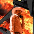 A low-quality photo of a man in a cowboy hat and light-colored jacket standing in front of a structure engulfed in bright orange flames.