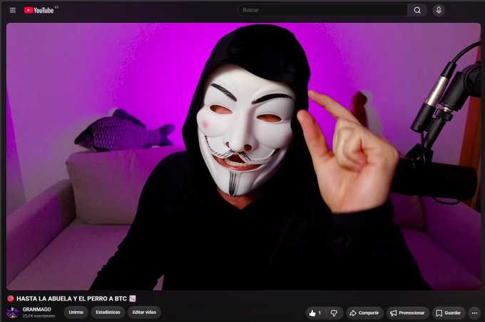 A YouTube thumbnail shows a person in a black hoodie wearing a white Guy Fawkes/V for Vendetta mask, pointing to their head against a pink-lit background with a microphone visible. The video titled "HASTA LA ABUELA Y EL PERRO A BTC" by GRANMAGO (with 25.8K subscribers) relates to the post's BTC update warning viewers not to get confident despite the FED sounding alarms.