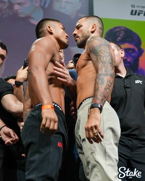 Two muscular, shirtless MMA fighters stand nose-to-nose in an intense staredown at a UFC weigh-in event, with one fighter's heavily tattooed arm prominently visible. The image relates to the post about UFC 331's "card built for LA," as the promotional banner in the background and the "Stake" sponsor logo hint at the upcoming September 19 fight event on Paramount Plus.