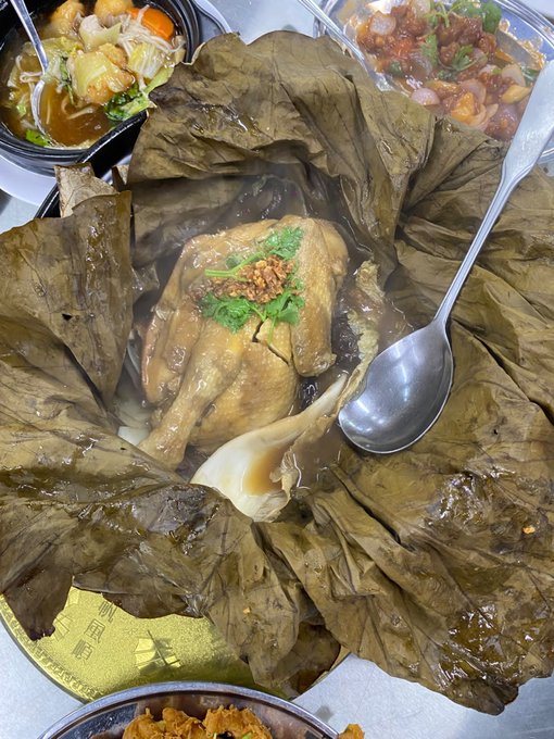 A traditional Chinese meal featuring a whole chicken cooked and served in a lotus leaf, accompanied by side dishes of vegetables and meat.