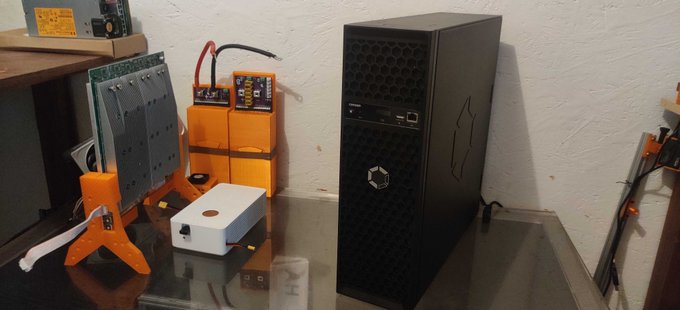 A black desktop computer tower sits on a table next to orange 3D-printed stands, circuit boards, and a small white electronic device.