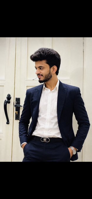 A young man with dark hair and a beard wears a navy blue suit with a white dress shirt and a belt featuring a distinctive buckle, standing in front of a white door.