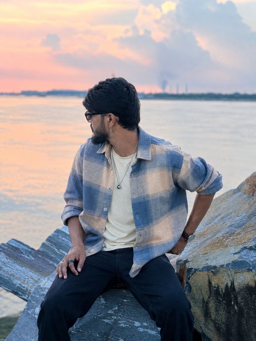 A young man with dark hair and glasses, wearing a blue and white plaid shirt over a cream t-shirt, sits on rocks by a calm body of water at sunset.