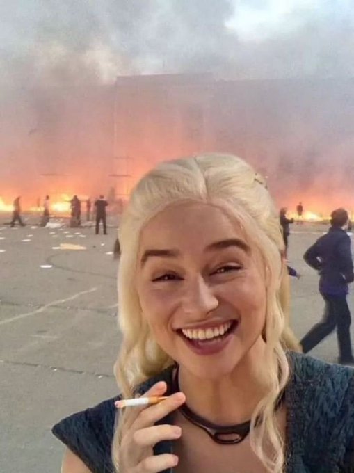 A blonde woman in a denim outfit smiles and holds a lit cigarette, taking a selfie in front of a large, smoky fire with people standing nearby.