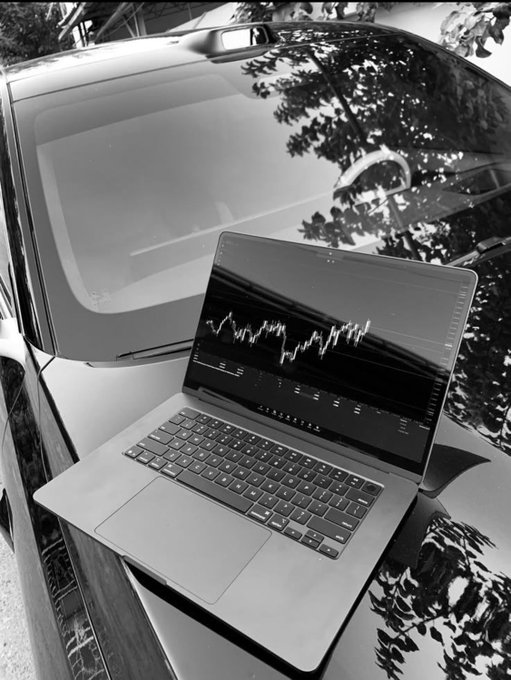 A black-and-white photo of an open laptop displaying a stock market chart resting on the hood of a reflective car.