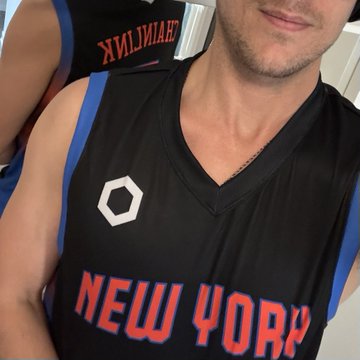 The image features a man wearing a black New York jersey with red lettering, standing against a white background. The man appears to be posing for a photo or selfie, as indicated by his smile. The caption of the image reads "Kicks win while in NYC for @ETHGlobal #7 @chainlink $LINK". This suggests that the man is wearing the New York jersey during an event at the New York Jets' stadium and has just won a game there.