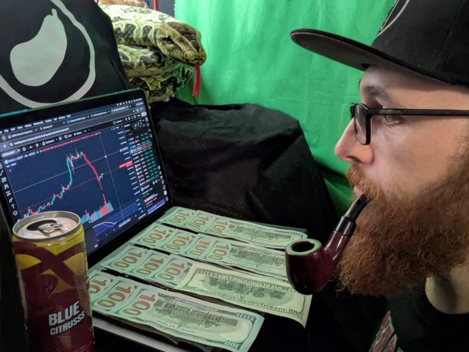 A man with a red beard, sunglasses, and a cap smoking a pipe while looking at a cryptocurrency trading chart on a laptop, with stacks of $100 bills and a Blue Citrus energy drink on the desk.
