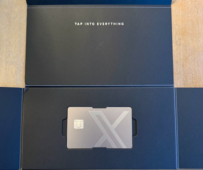 A dark navy presentation box opens to reveal a sleek black cardholder/wallet with an 'X' design, embossed with 'TAP INTO EVERYTHING' on the inner lid.