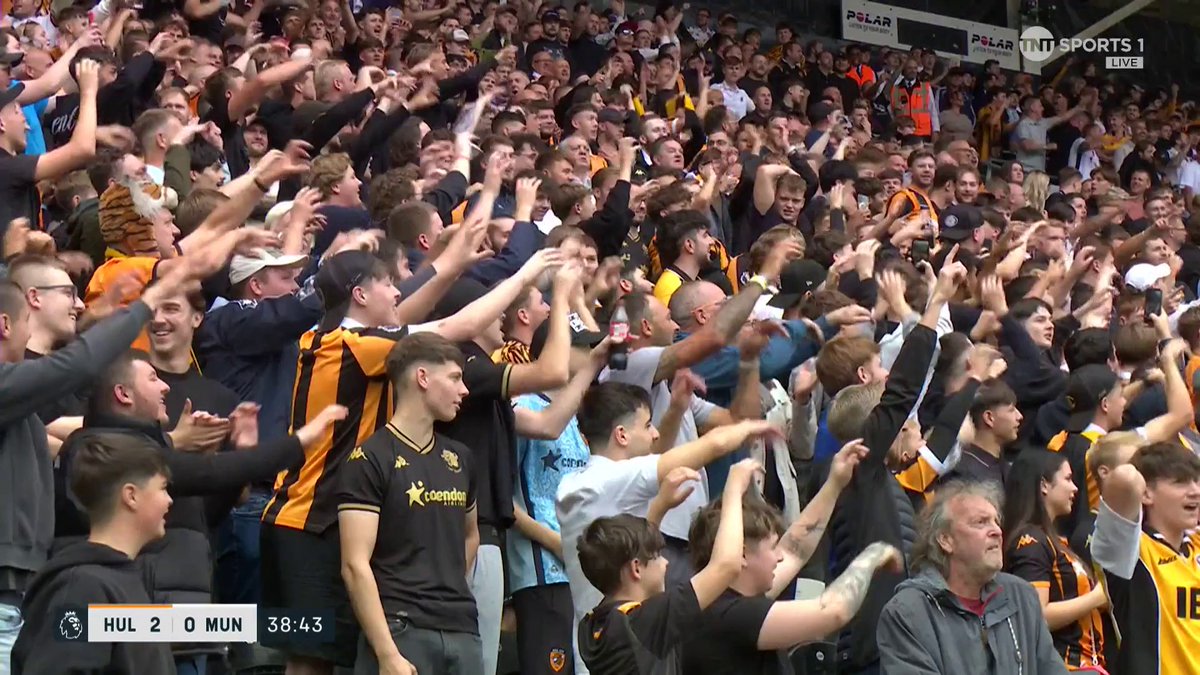 Hull City fans celebrate enthusiastically in the stands at the MKM Stadium, with the TNT Sports broadcast showing a 2-0 lead over Manchester United at the 38:43 mark, reflecting their delight at their team's dominant performance against the Red Devils.