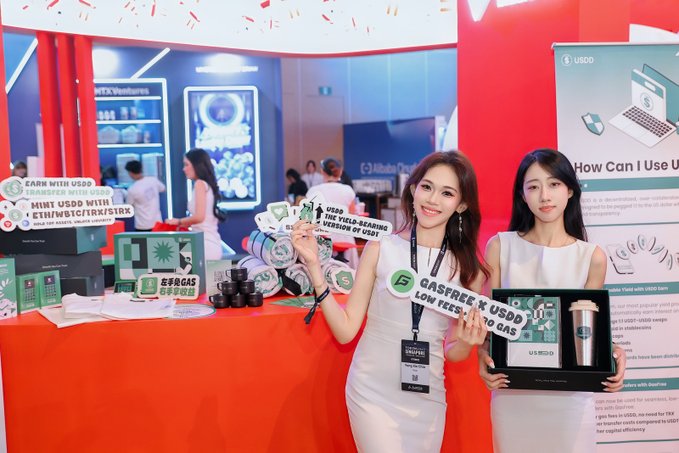 Two women stand at a trade show booth for USDD cryptocurrency, holding promotional signs; one sign reads 'GAS FREE X USDD LOW FEES TO GAS' and another says 'USDD THE YIELD-BEARING STABLECOIN BY USDD'.