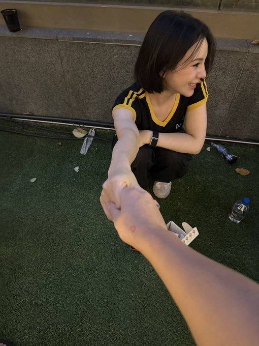 A person crouches on artificial turf, smiling and extending their hand toward the camera in what appears to be a fist bump or fist-bumping handshake.