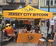 A vendor booth with a yellow canopy tent labeled 'JERSEY CITY BITCOIN' set up at an outdoor market, featuring a Bitcoin logo, merchandise displays, and a person wearing a Bitcoin-themed hat.
