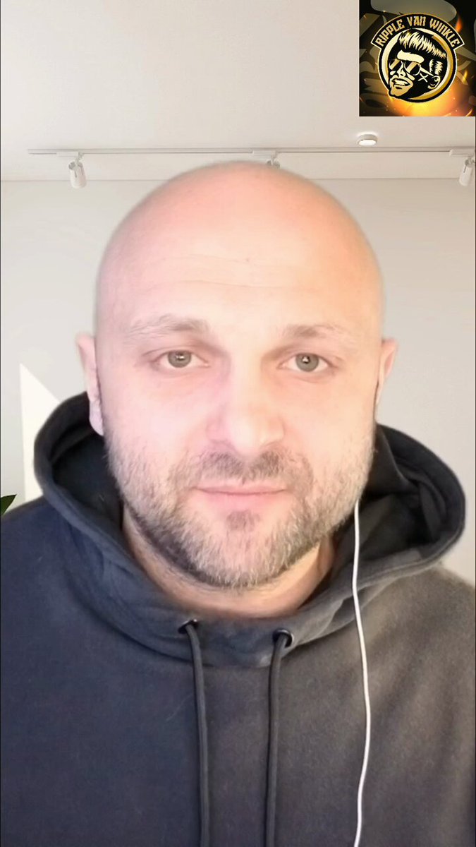 The image features a bald man wearing a black hoodie, with headphones around his neck. The background of the photo shows a white wall, which has an advertisement for a cryptocurrency exchange called "DTCC" displayed above it. The caption on the image reads "Focus on what is in the image and how it relates to the post".