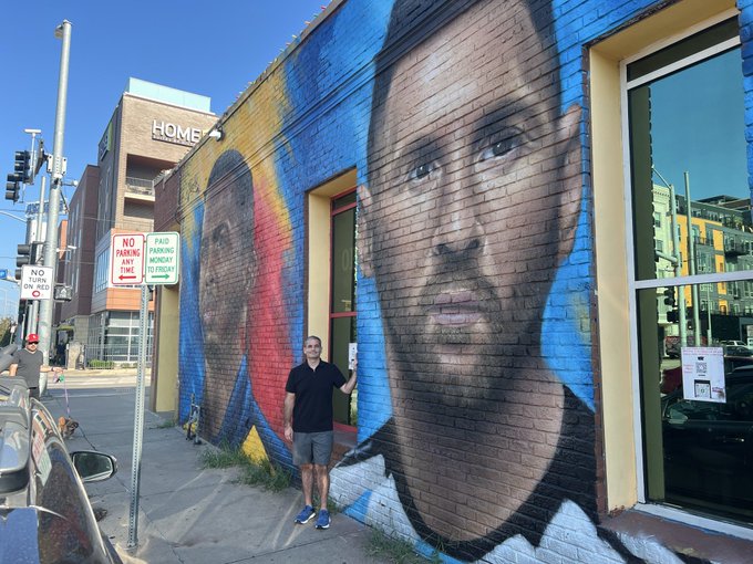 A man posing in front of a colorful outdoor mural on a building wall featuring large painted portraits of faces, with 'HOME' visible on an adjacent building and parking signs nearby.