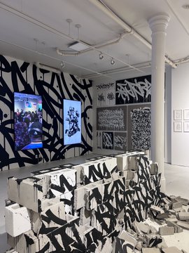 An art gallery space featuring black and white abstract murals and sculptures by artist RETNA, with two video screens displaying digital art.